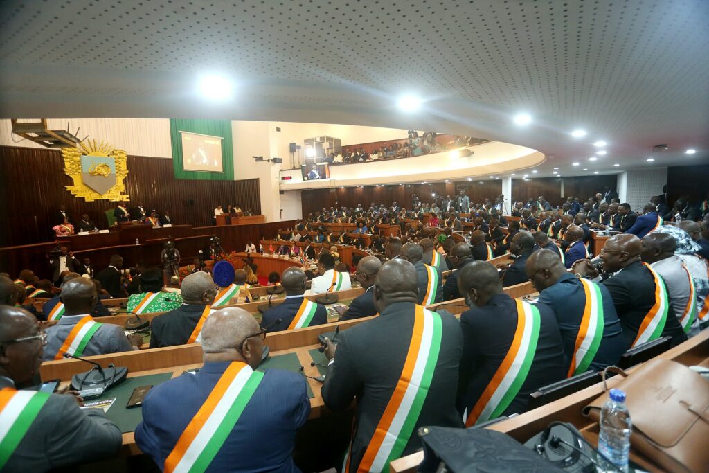 Hémicycle de l’Assemblée nationale ivoirienne lors de la rentrée parlementaire, députés portant l’écharpe orange, blanc, vert, armoiries au mur