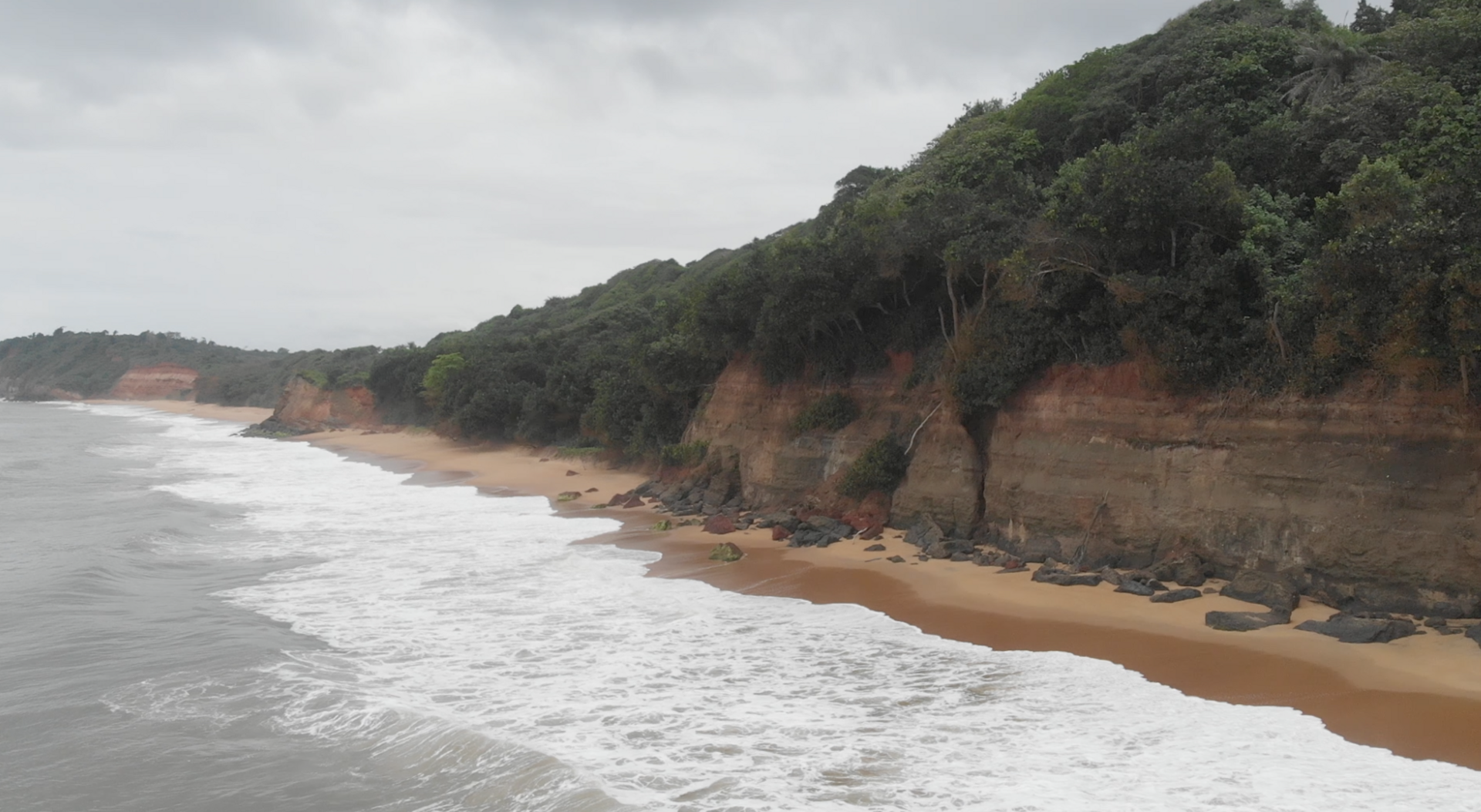 Falaises de terre rouge couvertes de végétation au-dessus d’une plage de sable battue par les vagues, à Fresco