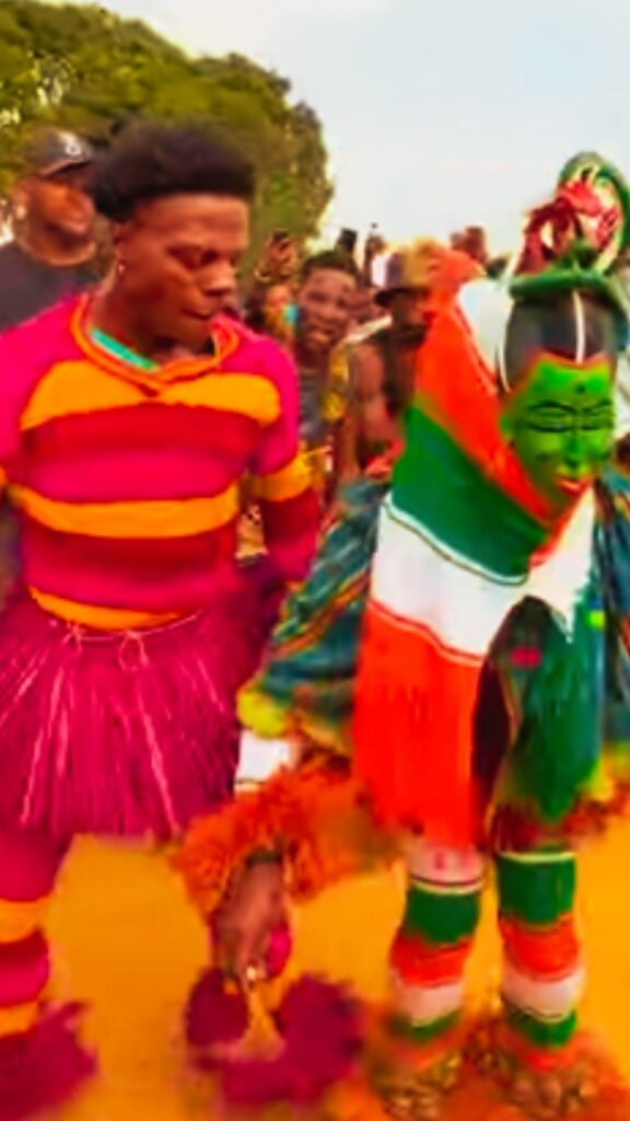 IShowSpeed, en tenue rose et orange à franges, danse aux côtés d'un danseur masqué au visage vert, devant la foule