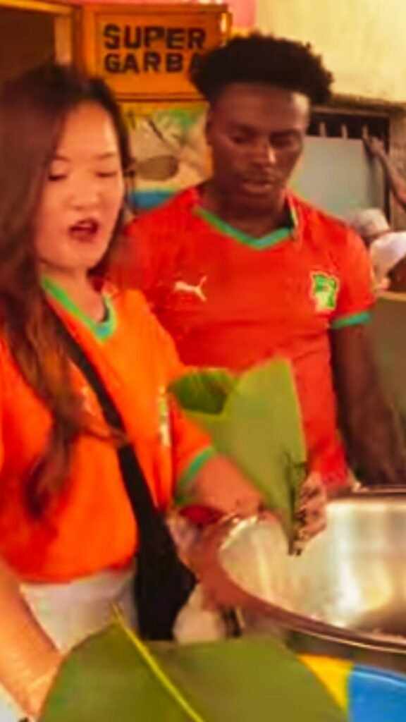 IShowSpeed en maillot orange des Éléphants derrière le comptoir d'un étal de garba, sous l'enseigne Super Garba