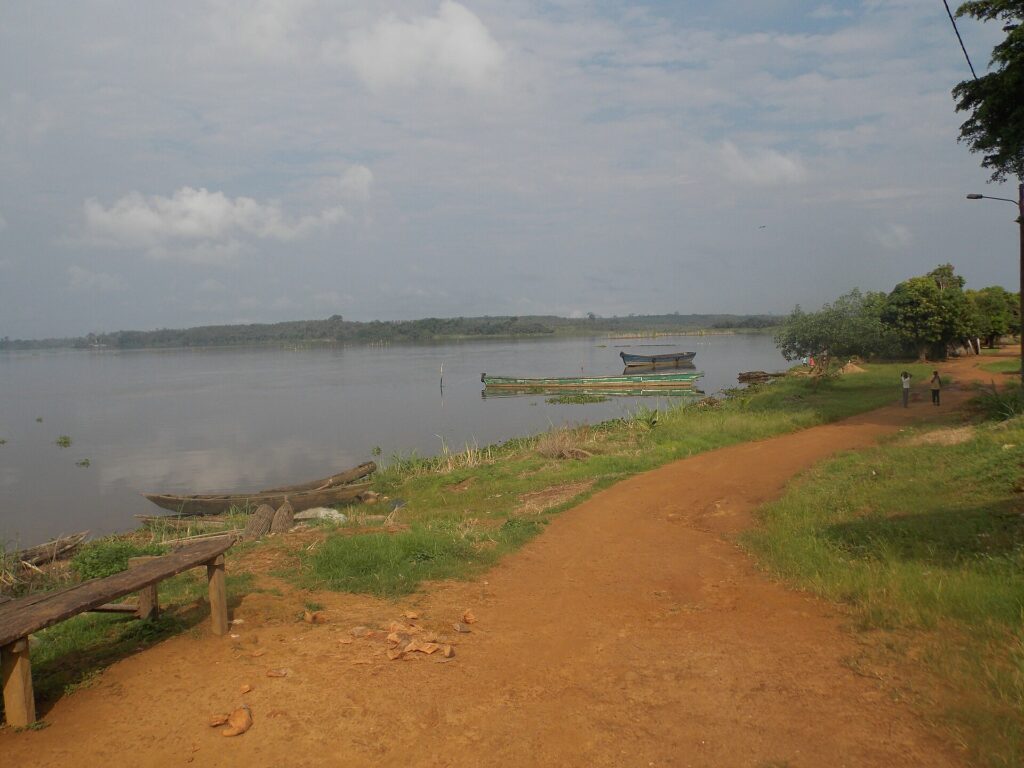 Rive de la lagune Aby avec un sentier de terre rouge, quelques pirogues et l’eau calme sous un ciel nuageux