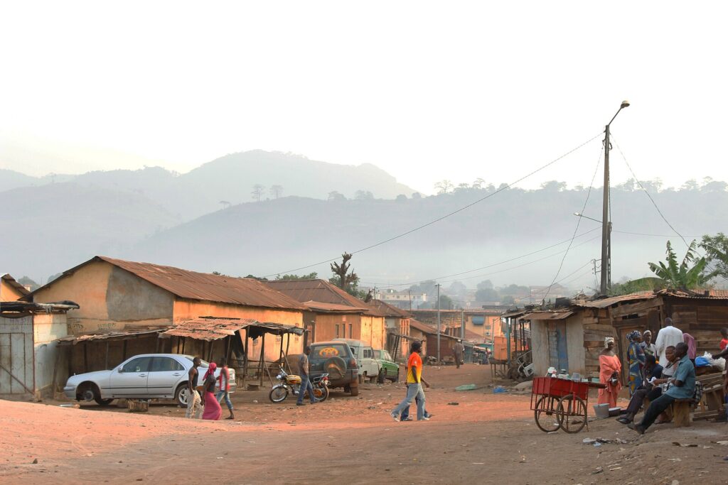 Rue en terre d’un quartier de Man, avec maisons basses et collines voilées de brume à l’arrière-plan