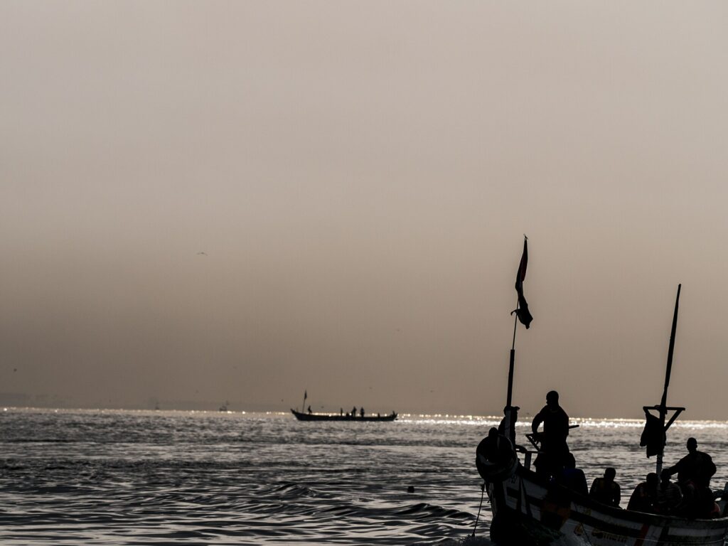 Silhouette de pêcheurs dans une pirogue au petit matin, sous un ciel voilé par la brume de l’harmattan, près de la côte ivoirienne